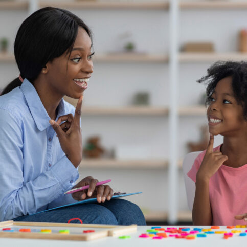 Speech therapist pretty young african american woman working with little black girl with bushy hair at clinic, sitting in front of each other, smiling and touching their chins, side view
