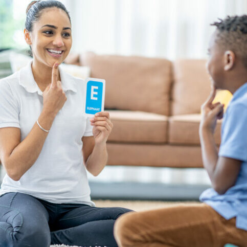 A female speech therapist sits with a young boy on the floor as they work on forming sounds.  She is holding up alphabet flash cards as they work together to make the sounds.